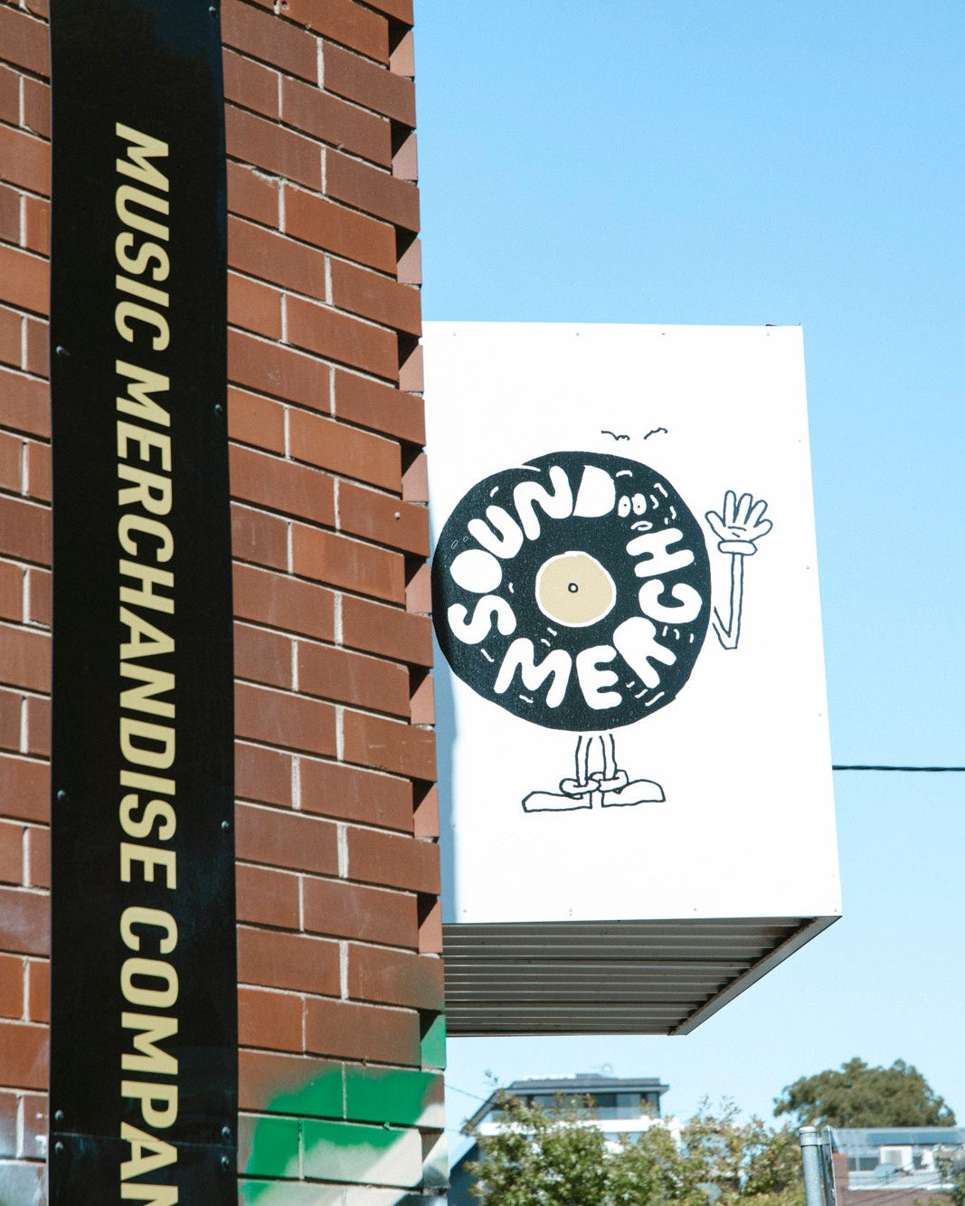 A white rectangular sign for Sound Merchant hanging from a brick building, featuring a black vinyl record logo with a small illustrated figure. A black vertical sign on the brick wall reads Music Merchandise Company in gold lettering.