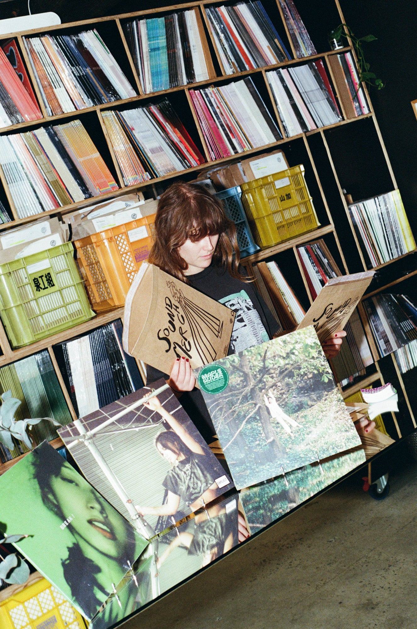 A person with shoulder-length brown hair stands in a record store or library area surrounded by vinyl records on shelves, holding two large vinyl record sleeves. One sleeve features a tan background with black script text, and the other shows a forest scene.